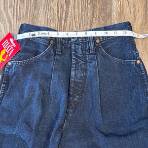 May include: Close-up of dark blue denim jeans with a high waist. A measuring tape shows the waist size. The jeans have pockets and a Wrangler RIATA tag.