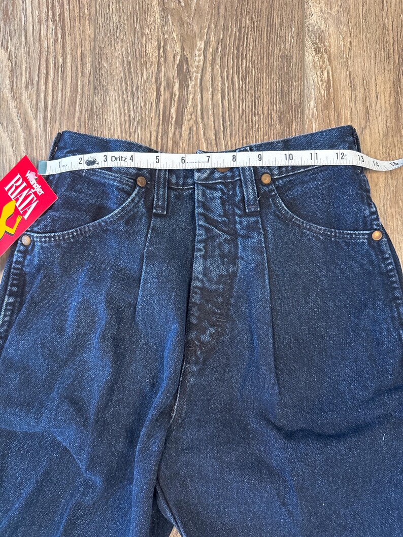 May include: Close-up of dark blue denim jeans with a high waist. A measuring tape shows the waist size. The jeans have pockets and a Wrangler RIATA tag.
