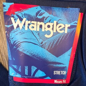 K&ouml;nnte beinhalten: Eine blaue Jeans aus Denim mit einem roten und wei&szlig;en Etikett mit der Aufschrift "Wrangler Stretch Misses Fit" und einem Bild einer Person, die Jeans tr&auml;gt.
