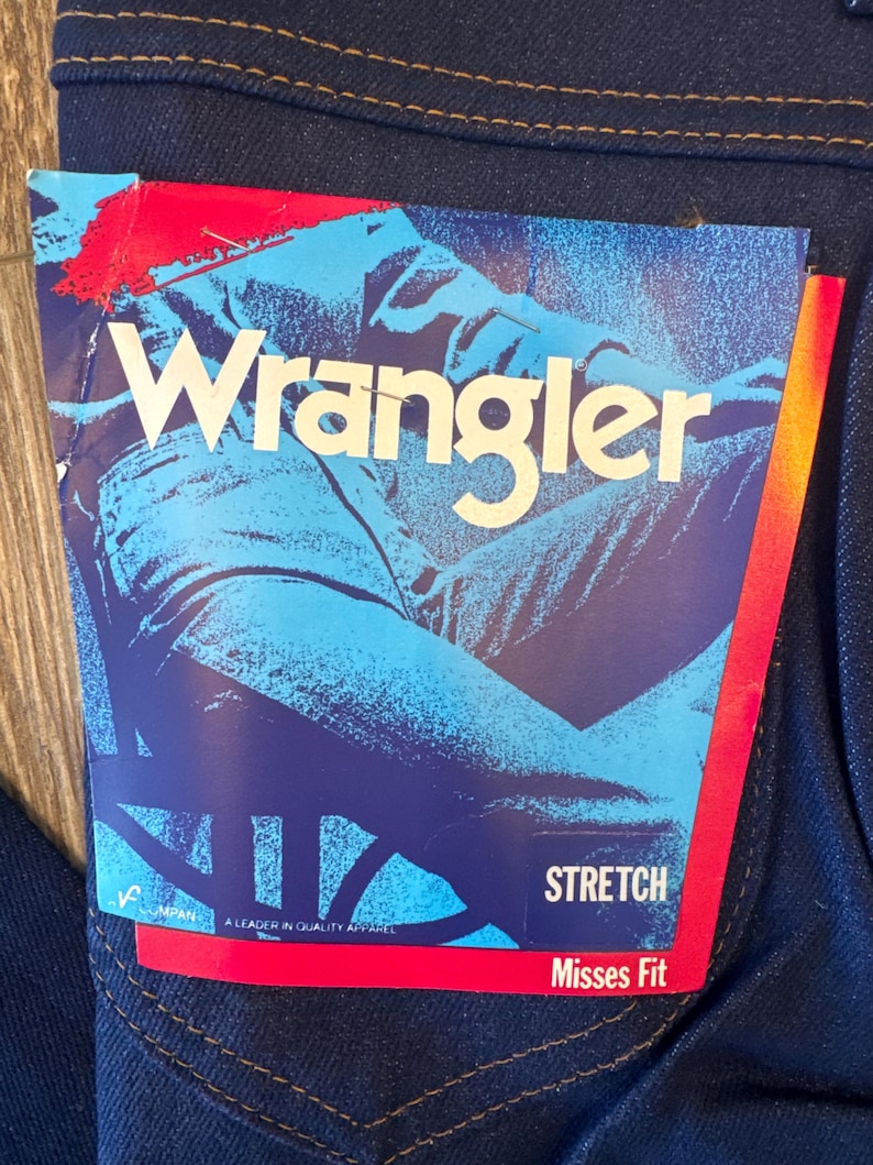 K&ouml;nnte beinhalten: Eine blaue Jeans aus Denim mit einem roten und wei&szlig;en Etikett mit der Aufschrift "Wrangler Stretch Misses Fit" und einem Bild einer Person, die Jeans tr&auml;gt.