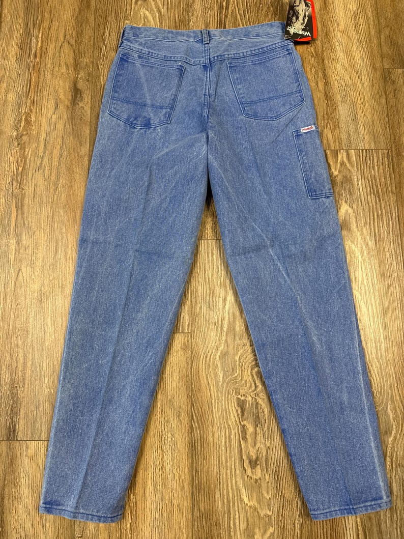 May include: Light blue denim jeans with two back pockets and a small side pocket. The jeans have a classic five-pocket design and a straight leg fit. The Wrangler tag is visible.