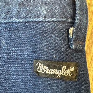 Puede incluir: Primer plano de una secci&oacute;n de vaqueros azul oscuro. Una etiqueta rectangular negra con la palabra "Wrangler" en escritura blanca est&aacute; cosida a los vaqueros. El denim tiene un aspecto texturizado.