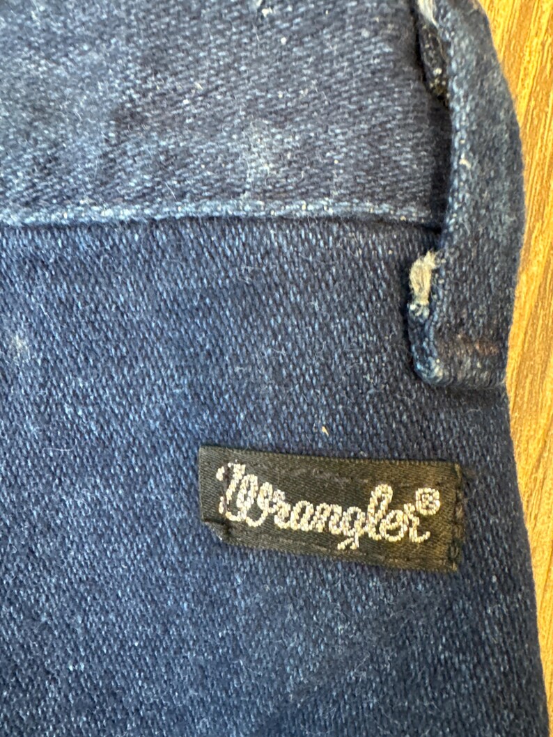 Puede incluir: Primer plano de una secci&oacute;n de vaqueros azul oscuro. Una etiqueta rectangular negra con la palabra "Wrangler" en escritura blanca est&aacute; cosida a los vaqueros. El denim tiene un aspecto texturizado.