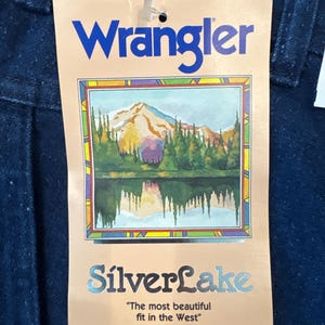 Puede incluir: Primer plano de una etiqueta de jeans Wrangler. La etiqueta es de color beige con la palabra "Wrangler" en azul. La etiqueta tiene una ilustraci&oacute;n de una cordillera y un lago. Las palabras "SilverLake" y "The most beautiful fit in the West" tambi&eacute;n est&aacute;n en la etiqueta.