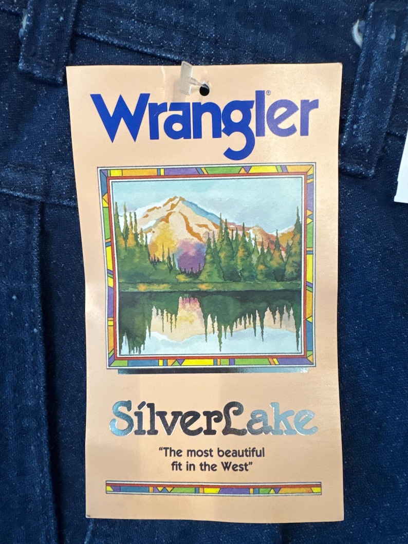 Puede incluir: Primer plano de una etiqueta de jeans Wrangler. La etiqueta es de color beige con la palabra "Wrangler" en azul. La etiqueta tiene una ilustraci&oacute;n de una cordillera y un lago. Las palabras "SilverLake" y "The most beautiful fit in the West" tambi&eacute;n est&aacute;n en la etiqueta.