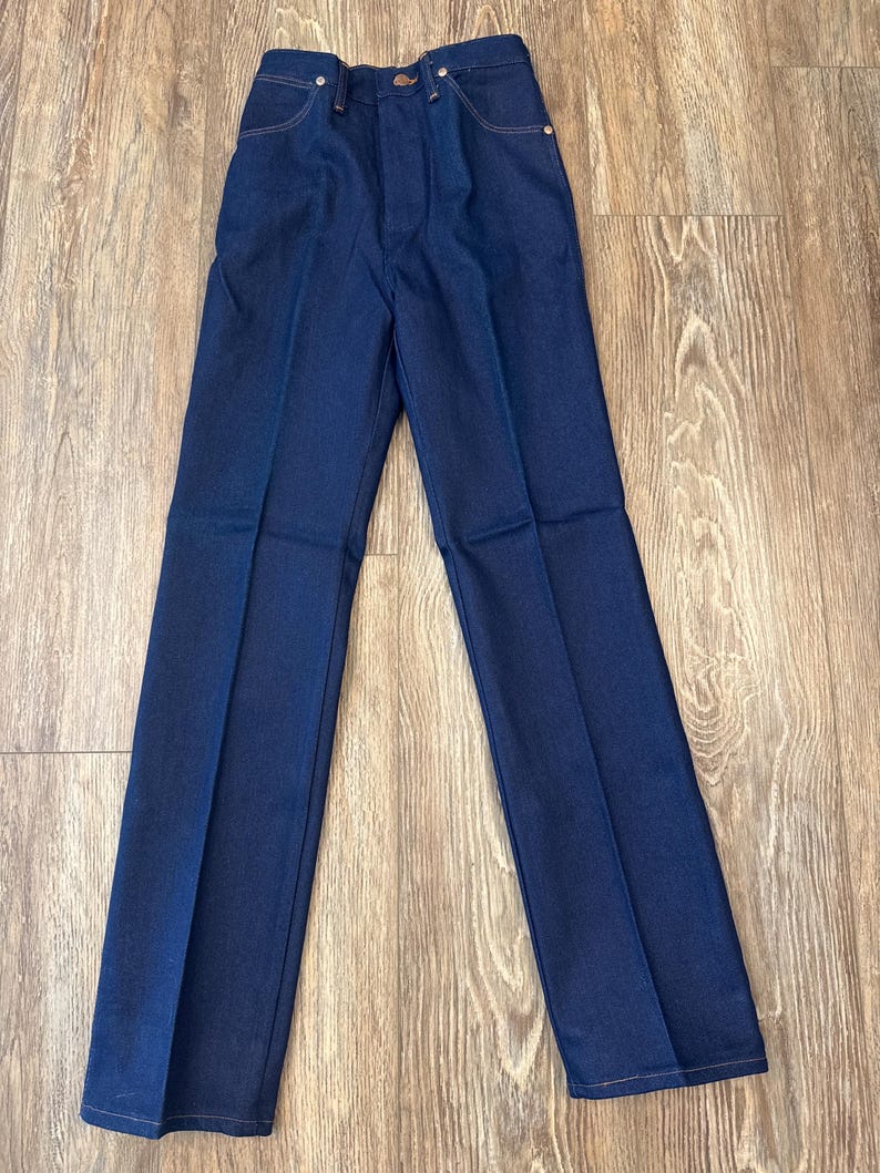 May include: Dark blue denim pants with a classic five-pocket design. The pants have a button and zipper closure, with a straight leg cut. The stitching is a contrasting light brown color. The pants are laid flat on a wooden surface.