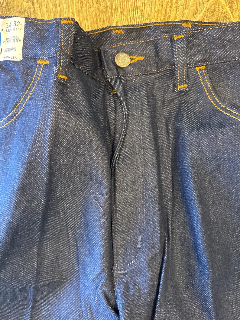 Peut inclure: Un jean en denim bleu fonc&eacute; avec un bouton marron et des coutures orange. Le jean porte une &eacute;tiquette de taille indiquant "34 32" et "8411PS".