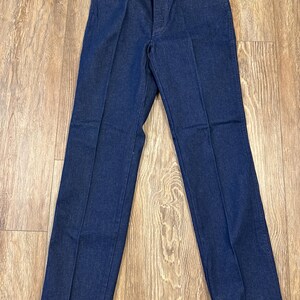 May include: Dark blue denim jeans with a straight leg cut. The jeans have a classic five-pocket design, a button and zipper closure, and belt loops. A tag with the brand name "Wrangler" is attached to the jeans.
