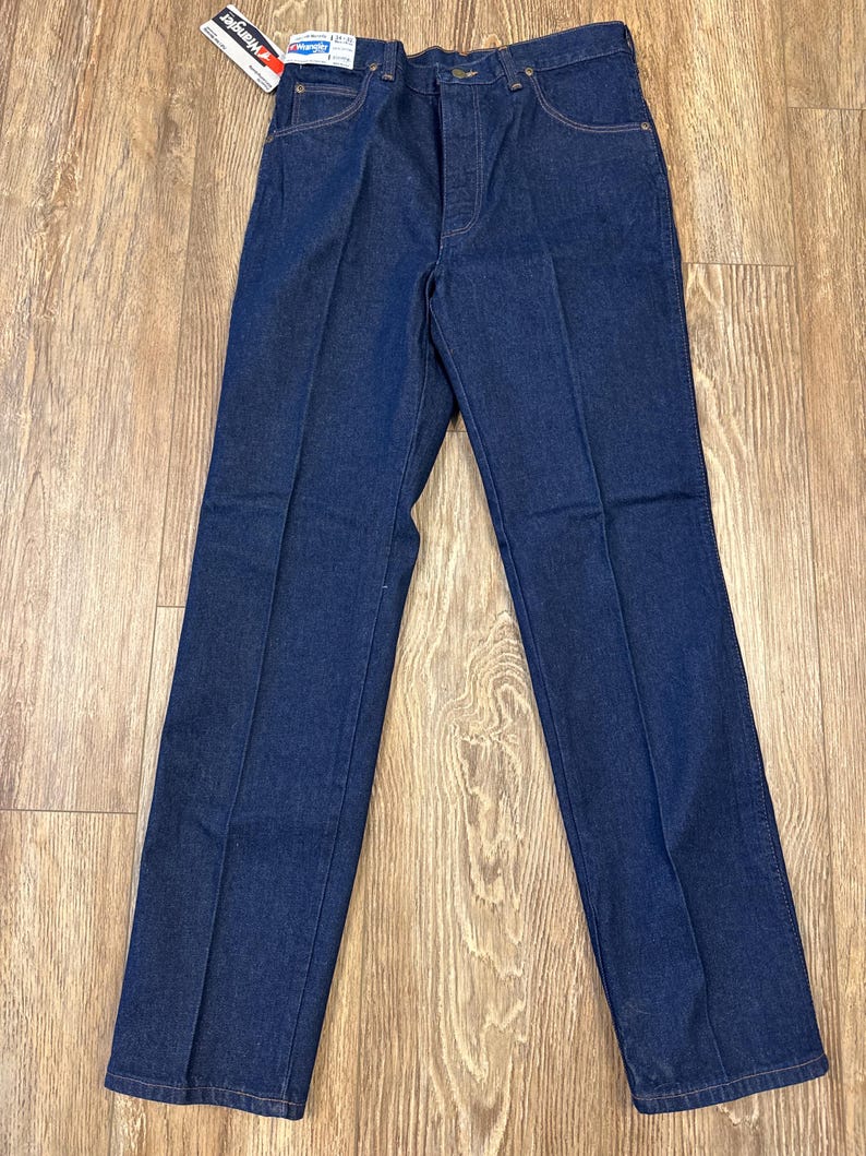 May include: Dark blue denim jeans with a straight leg cut. The jeans have a classic five-pocket design, a button and zipper closure, and belt loops. A tag with the brand name "Wrangler" is attached to the jeans.