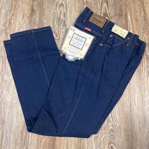 May include: Dark blue Wrangler Classic Jeans, size 28x34, with a paper tag attached. The jeans are folded and laid on a wooden surface. The Wrangler logo is visible on the back pocket and a leather patch.