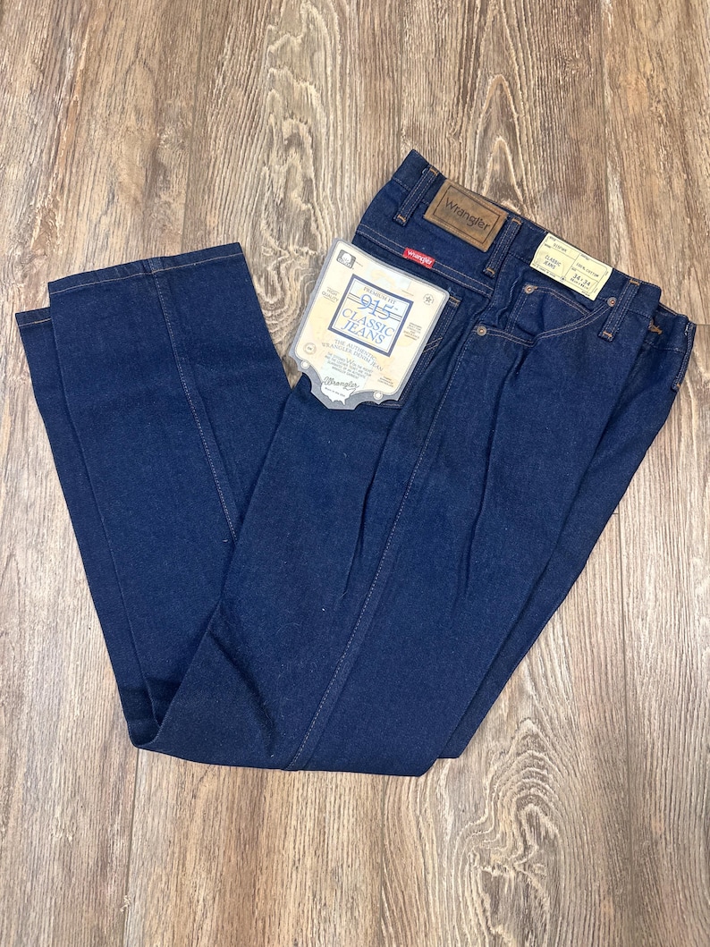 May include: Dark blue Wrangler Classic Jeans, size 28x34, with a paper tag attached. The jeans are folded and laid on a wooden surface. The Wrangler logo is visible on the back pocket and a leather patch.