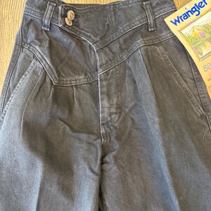 May include: Close-up of a pair of dark grey Wrangler jeans. The jeans have a high waist, pleats, and a unique button closure. A Wrangler tag is visible on the right side. The jeans are made of a durable denim material.