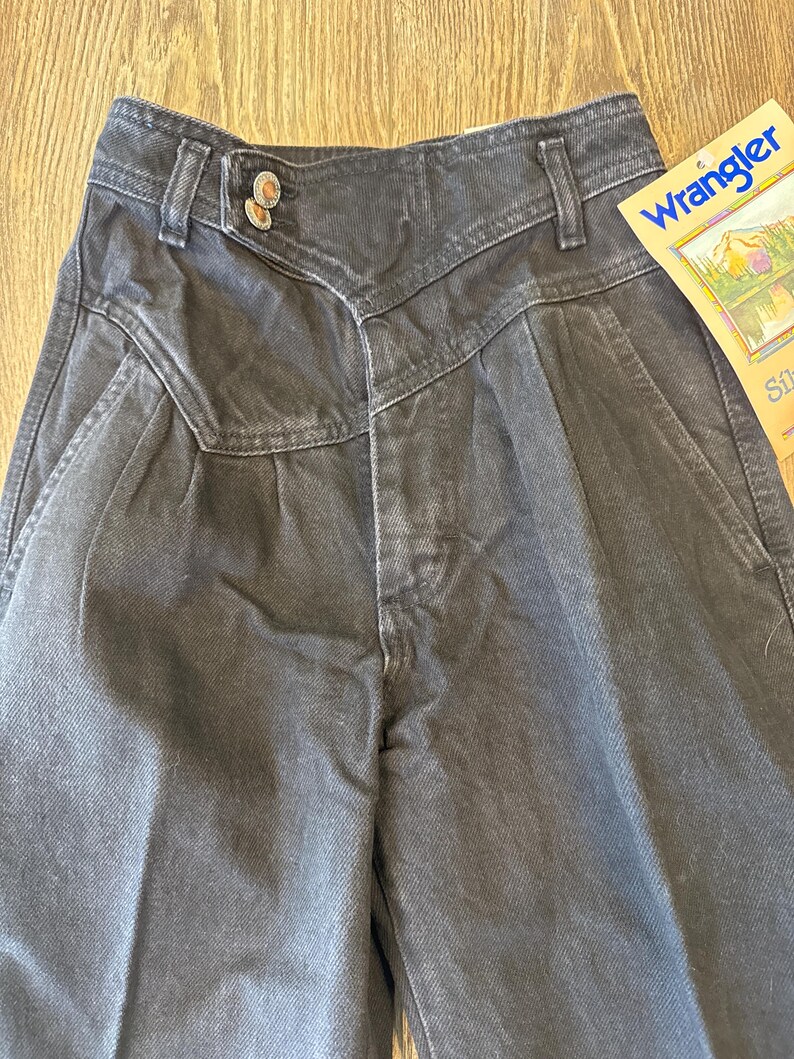 May include: Close-up of a pair of dark grey Wrangler jeans. The jeans have a high waist, pleats, and a unique button closure. A Wrangler tag is visible on the right side. The jeans are made of a durable denim material.