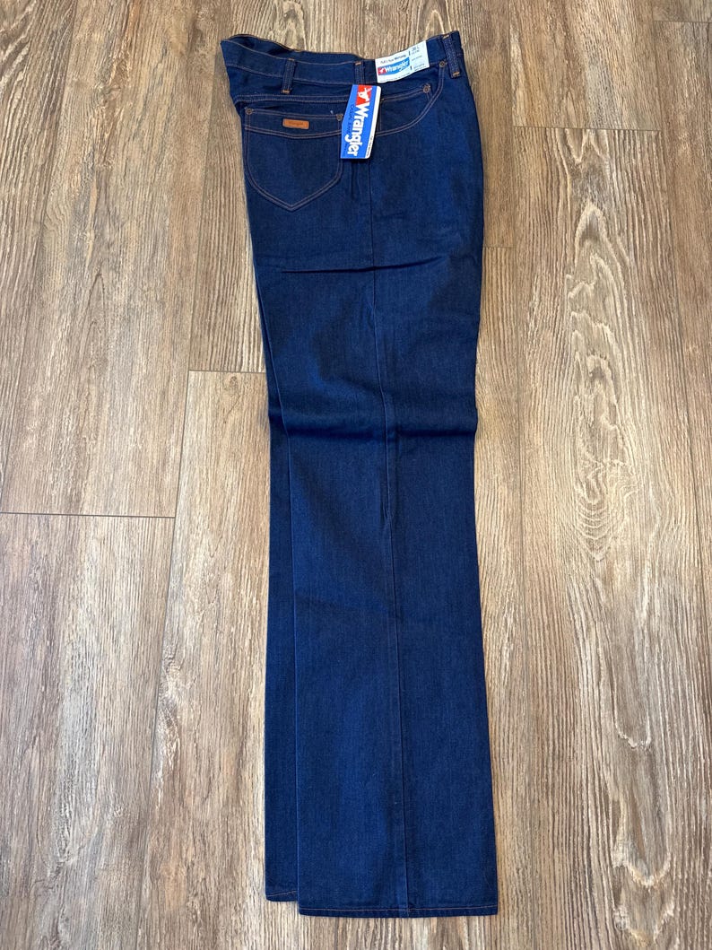 May include: A pair of dark blue denim jeans with a Wrangler brand tag attached. The jeans have a straight leg style and are folded at the bottom.