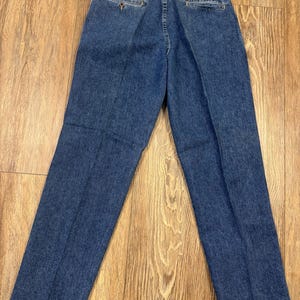 May include: Blue denim jeans, seen from the rear, featuring a button closure and pockets. The jeans have a classic, straight-leg cut and a small tag attached to the back pocket. The denim has a medium wash.
