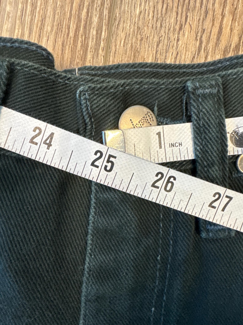 May include: Close-up of a dark green fabric with a white measuring tape that reads "24", "25", "26", "27", and "1 inch".