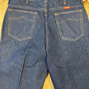 May include: Dark blue denim jeans with the Wrangler logo on the back pocket. The jeans have classic five-pocket styling with yellow stitching and a straight leg. The back pockets have a unique curved design.