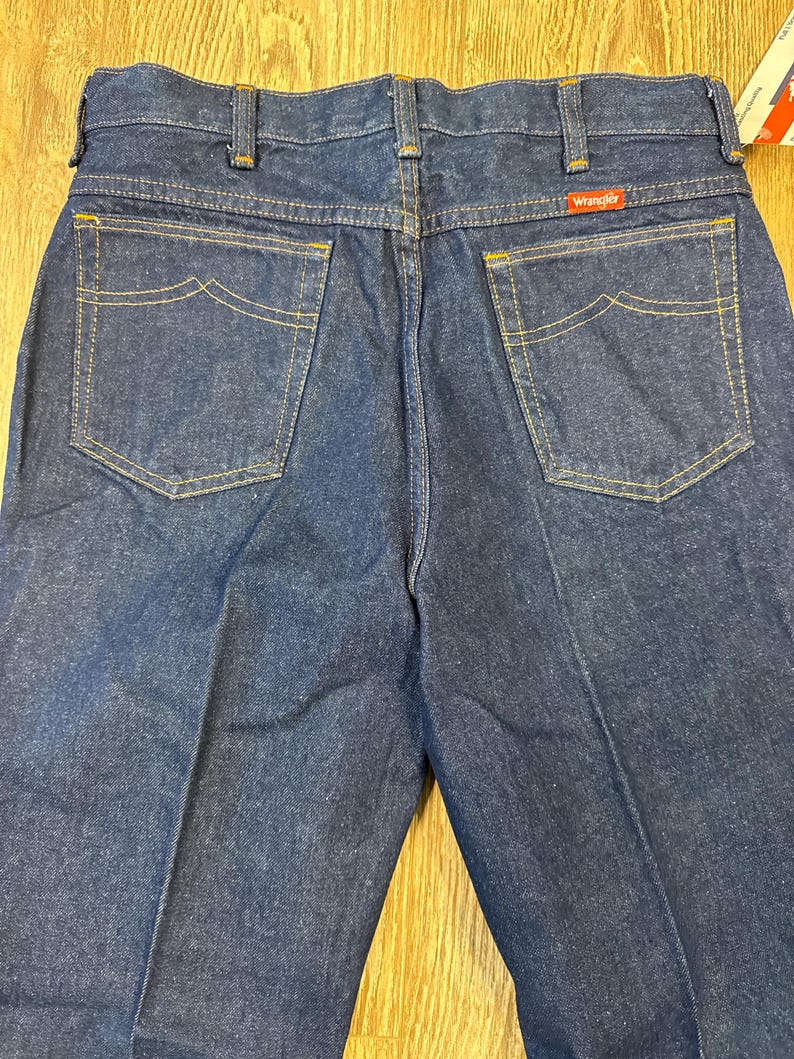May include: Dark blue denim jeans with the Wrangler logo on the back pocket. The jeans have classic five-pocket styling with yellow stitching and a straight leg. The back pockets have a unique curved design.