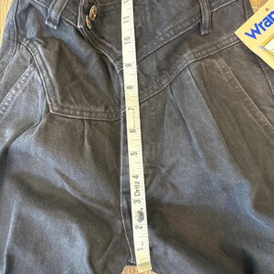 May include: Close-up of black denim Wrangler jeans with a high waist and pleated front. A measuring tape shows a 30.5 cm inseam. The jeans have a button closure and a Wrangler tag.