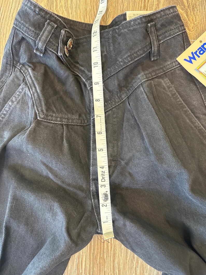 May include: Close-up of black denim Wrangler jeans with a high waist and pleated front. A measuring tape shows a 30.5 cm inseam. The jeans have a button closure and a Wrangler tag.