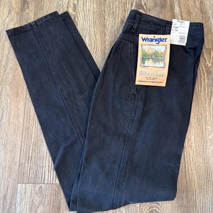 May include: Dark wash Wrangler Silverlake jeans with a high waist and a straight leg. The jeans have a tag attached with the Wrangler logo and a scenic image. The pants are folded on a wooden surface.