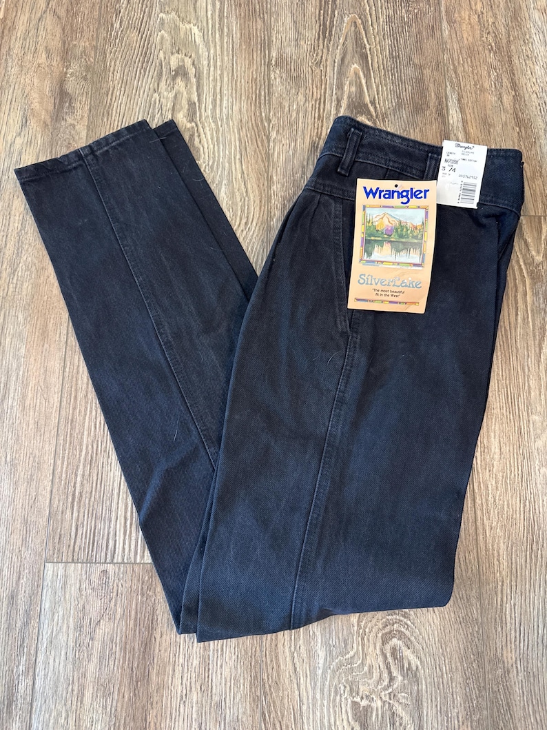 May include: Dark wash Wrangler Silverlake jeans with a high waist and a straight leg. The jeans have a tag attached with the Wrangler logo and a scenic image. The pants are folded on a wooden surface.