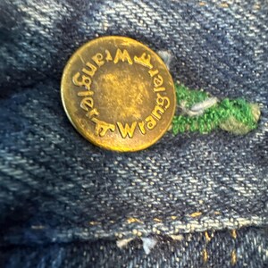 May include: Close-up of a brass button on blue denim fabric. The button is embossed with the word "Wrangler" in a circular pattern. Green stitching is visible near the button.