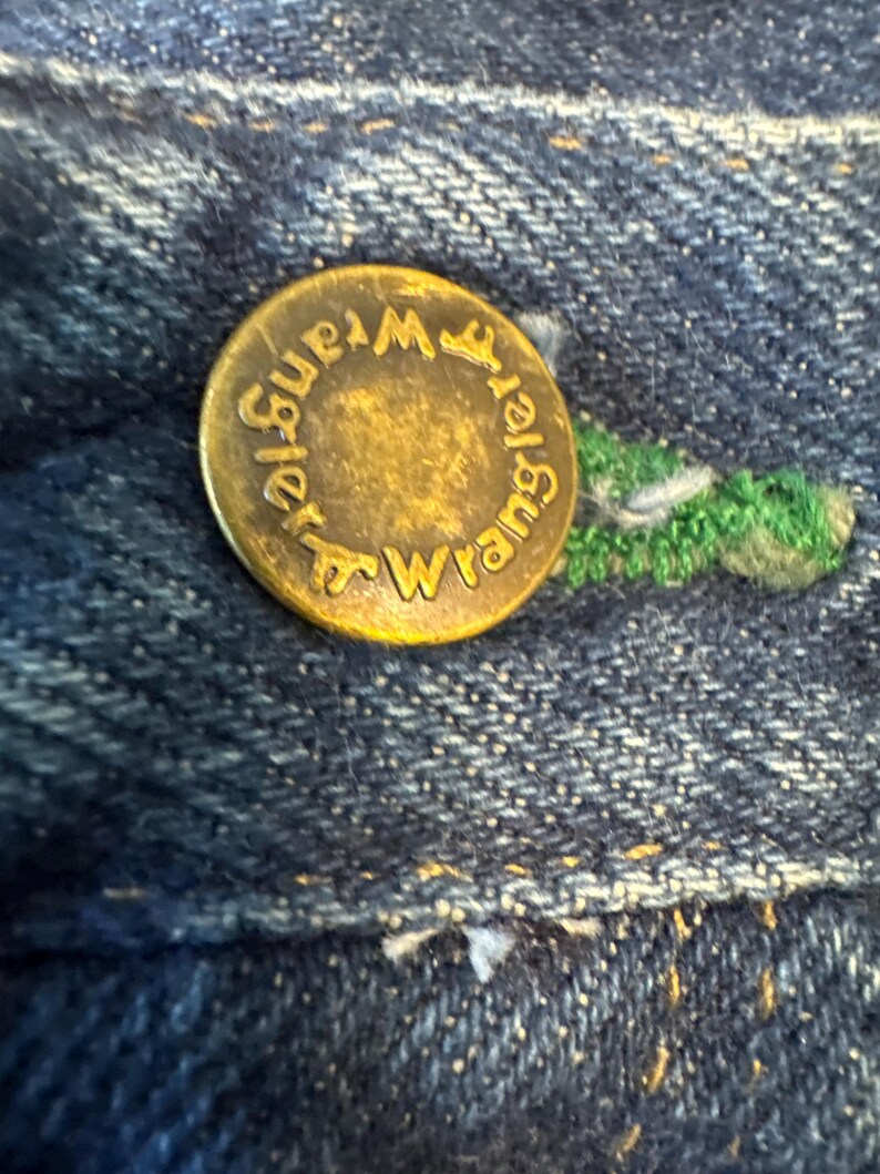May include: Close-up of a brass button on blue denim fabric. The button is embossed with the word "Wrangler" in a circular pattern. Green stitching is visible near the button.