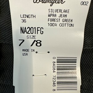 Op de afbeelding: Een wit label met zwarte tekst, een barcode en het Wrangler-logo. Het label vermeldt de productdetails: Silverlake WPRA Jean, Forest Green, 100% katoen, Lengte 36, Maat 7/8, NA201FG, Gemaakt in de VS, S474 002 en 0 84084 72346 8.