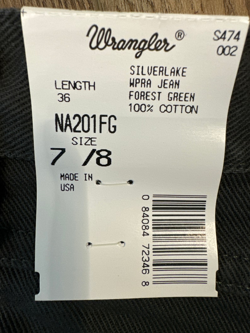 Op de afbeelding: Een wit label met zwarte tekst, een barcode en het Wrangler-logo. Het label vermeldt de productdetails: Silverlake WPRA Jean, Forest Green, 100% katoen, Lengte 36, Maat 7/8, NA201FG, Gemaakt in de VS, S474 002 en 0 84084 72346 8.