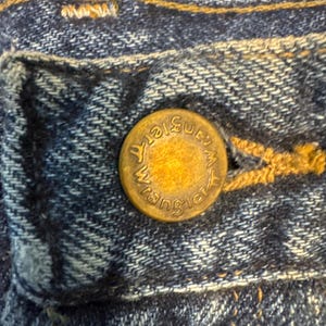 Puede incluir: Primer plano de una secci&oacute;n de vaqueros azules. Un bot&oacute;n de color lat&oacute;n con la palabra "Wrangler" est&aacute; centrado en la imagen. El bot&oacute;n est&aacute; sujeto a la zona del bolsillo de los vaqueros, con costuras visibles.