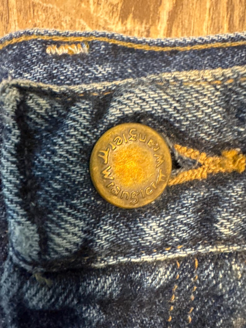 Puede incluir: Primer plano de una secci&oacute;n de vaqueros azules. Un bot&oacute;n de color lat&oacute;n con la palabra "Wrangler" est&aacute; centrado en la imagen. El bot&oacute;n est&aacute; sujeto a la zona del bolsillo de los vaqueros, con costuras visibles.