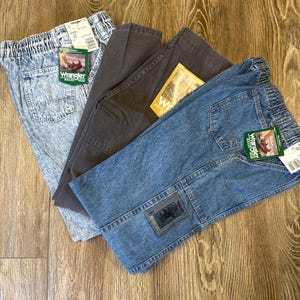 K&ouml;nnte beinhalten: Drei Jeanshosen von Wrangler Rugged Wear in Acid-Wash, Anthrazitgrau und blauem Denim. Jede Hose hat einen elastischen Bund und ein Wrangler-Etikett. Die blaue Jeans hat einen Aufn&auml;her am Knie.