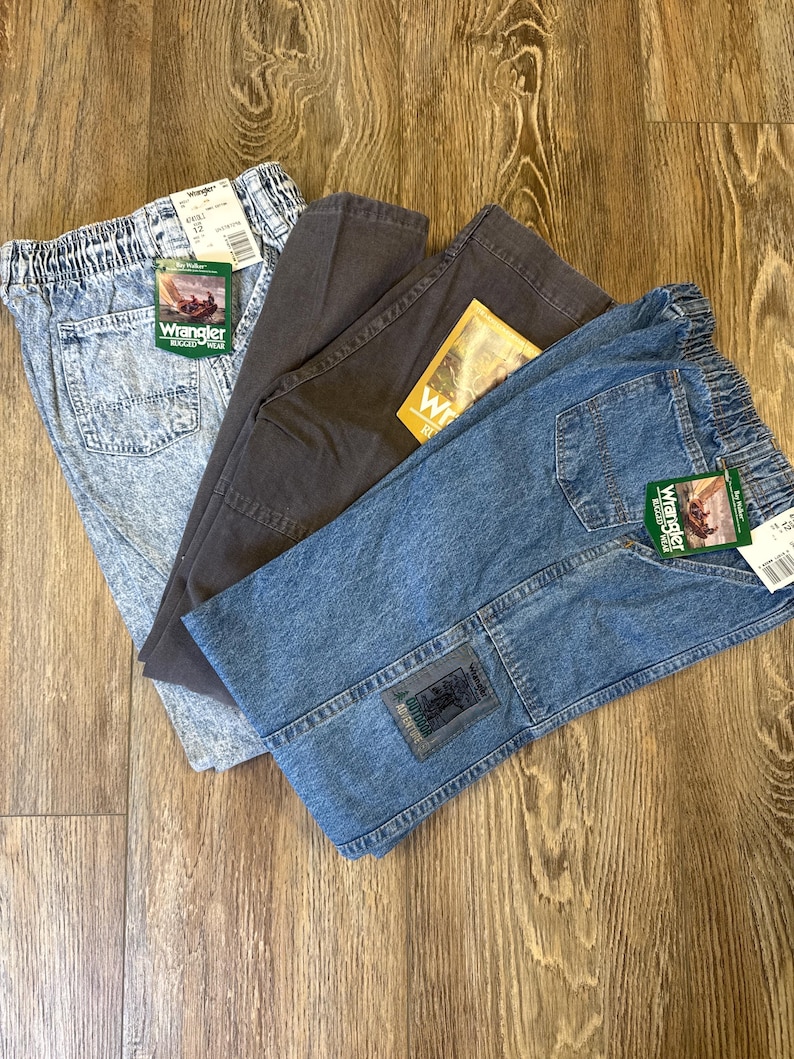 K&ouml;nnte beinhalten: Drei Jeanshosen von Wrangler Rugged Wear in Acid-Wash, Anthrazitgrau und blauem Denim. Jede Hose hat einen elastischen Bund und ein Wrangler-Etikett. Die blaue Jeans hat einen Aufn&auml;her am Knie.