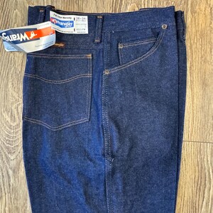 May include: Dark blue Wrangler jeans with a tag attached. The jeans have a classic five-pocket design with a straight leg. The tag reads "Wrangler" and includes size information: 38x34. The jeans are made of 100% cotton.
