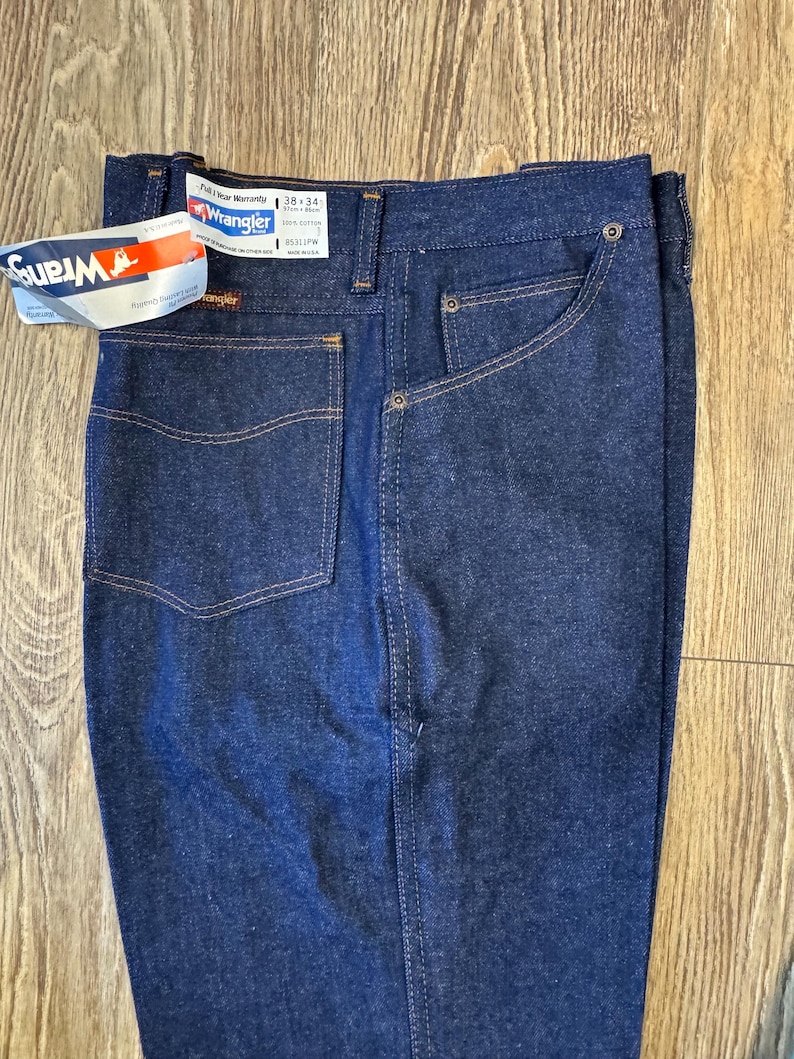 May include: Dark blue Wrangler jeans with a tag attached. The jeans have a classic five-pocket design with a straight leg. The tag reads "Wrangler" and includes size information: 38x34. The jeans are made of 100% cotton.