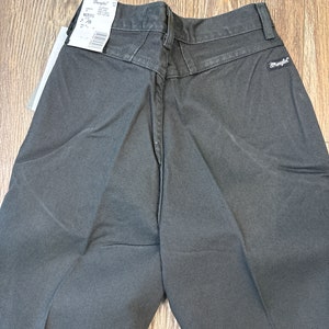 Op de afbeelding: Een donkergrijze Wrangler broek met een label aan de achterkant van de tailleband. Het label zegt "Wrangler" en heeft een maat van 32/32.