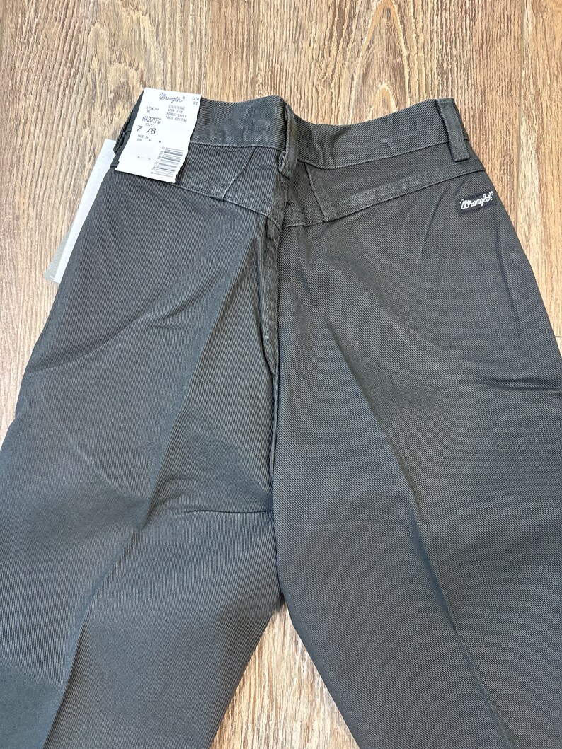 Op de afbeelding: Een donkergrijze Wrangler broek met een label aan de achterkant van de tailleband. Het label zegt "Wrangler" en heeft een maat van 32/32.
