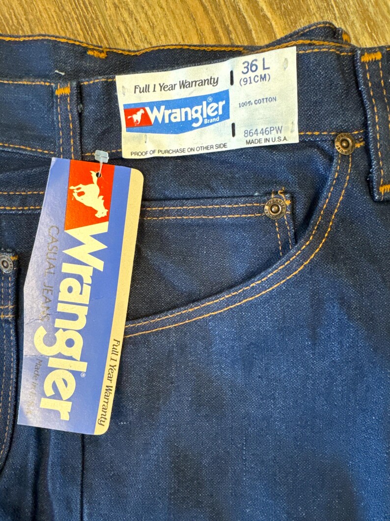 May include: A pair of blue denim jeans with a Wrangler brand tag. The tag says "Full 1 Year Warranty" and "100% Cotton". The size is 36 L (91 cm) and the style number is 86446PW. The jeans are made in the USA.