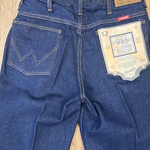May include: Close-up of dark blue Wrangler 915 Classic Jeans. The jeans have a classic five-pocket design with the Wrangler logo on the back pocket and a tag that reads "Premium Fit 915 Classic Jeans". The stitching is a contrasting gold color.