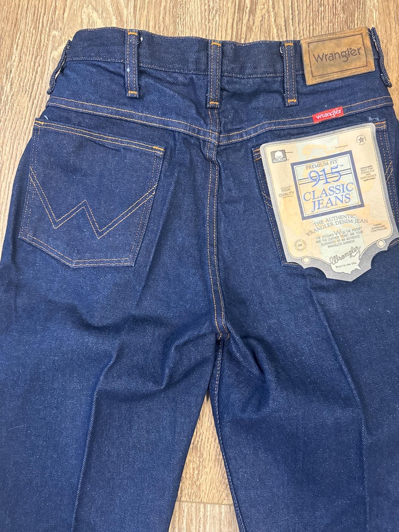 May include: Close-up of dark blue Wrangler 915 Classic Jeans. The jeans have a classic five-pocket design with the Wrangler logo on the back pocket and a tag that reads "Premium Fit 915 Classic Jeans". The stitching is a contrasting gold color.