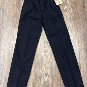 May include: Black denim Wrangler jeans with a straight leg and a high waist. The jeans have a button closure and a zipper fly. A tag with the Wrangler logo is attached to the jeans.
