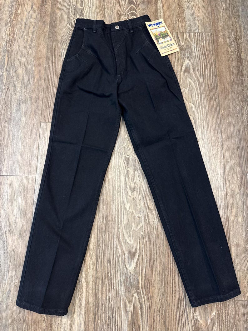 May include: Black denim Wrangler jeans with a straight leg and a high waist. The jeans have a button closure and a zipper fly. A tag with the Wrangler logo is attached to the jeans.