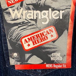 May include: Close-up of dark blue denim jeans with a Wrangler tag. The tag features the Wrangler logo, the words "AMERICAN HERO", and "STONE WASHED". The jeans have a classic five-pocket design.