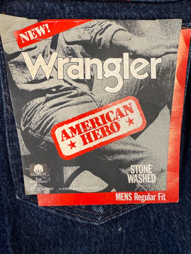 May include: Close-up of dark blue denim jeans with a Wrangler tag. The tag features the Wrangler logo, the words "AMERICAN HERO", and "STONE WASHED". The jeans have a classic five-pocket design.