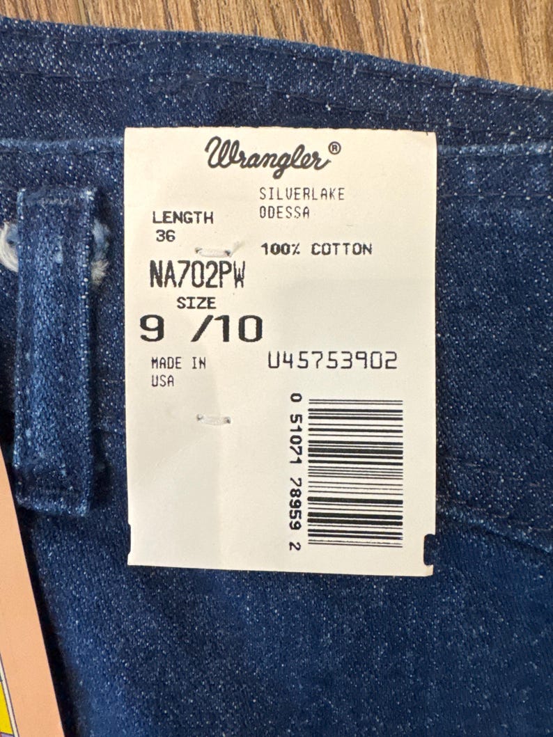 Puede incluir: Primer plano de tela vaquera azul oscuro con una etiqueta blanca adjunta. La etiqueta dice "Wrangler" e incluye detalles como "SILVERLAKE ODESSA", "100% COTTON", talla 9/10 y una longitud de 91 cm. La etiqueta tambi&eacute;n tiene un c&oacute;digo de barras.