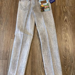May include: Light gray Wrangler Cowboy Cut jeans with a faded wash. The jeans have a classic five-pocket design, a Wrangler patch on the back pocket, and a tag that reads "Cowboy Cut". The jeans are displayed on a wooden surface.