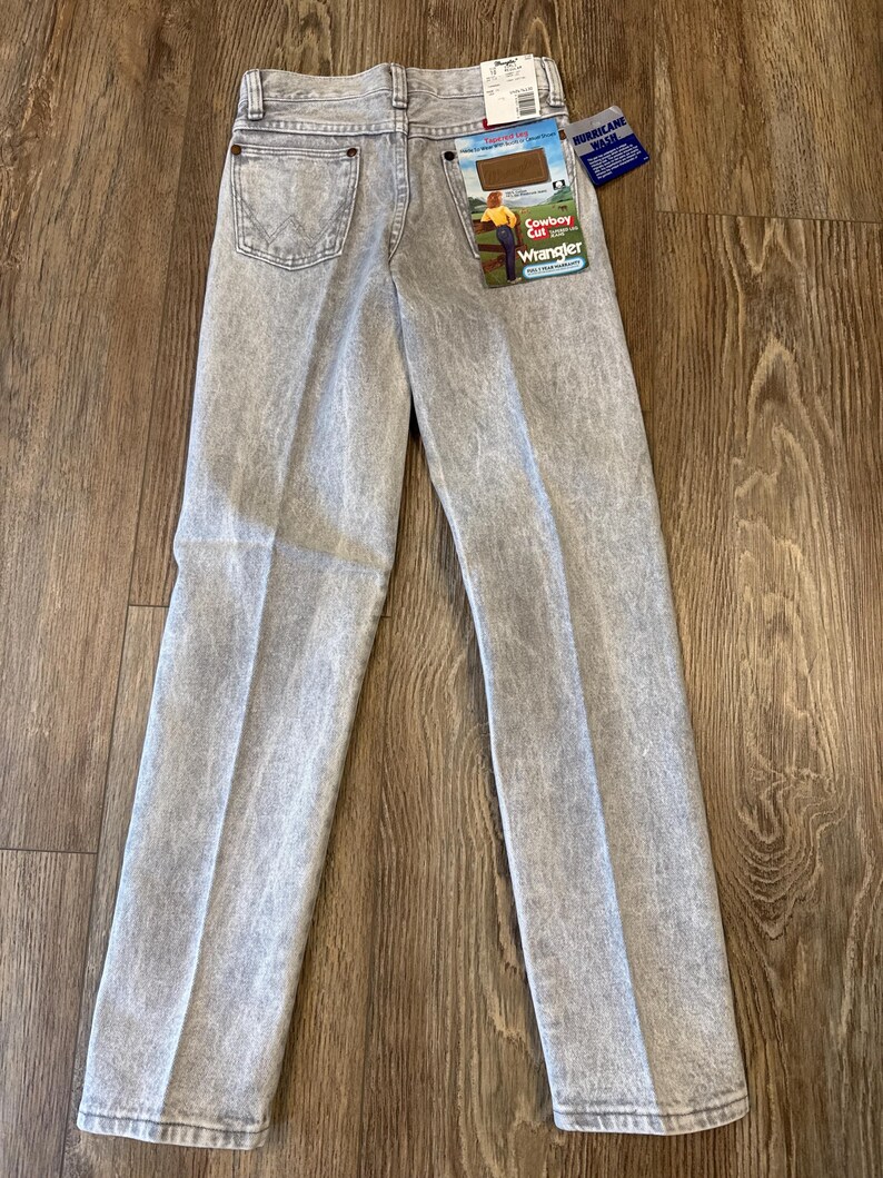 May include: Light gray Wrangler Cowboy Cut jeans with a faded wash. The jeans have a classic five-pocket design, a Wrangler patch on the back pocket, and a tag that reads "Cowboy Cut". The jeans are displayed on a wooden surface.