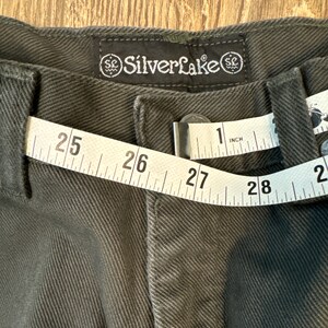 Op de afbeelding: Een donkergrijze broek met een meetlint om de taille. Het lint geeft 25 tot 28 inch aan. De broek heeft een label met de tekst "Silverlake" en een gestileerd "SC"-logo.
