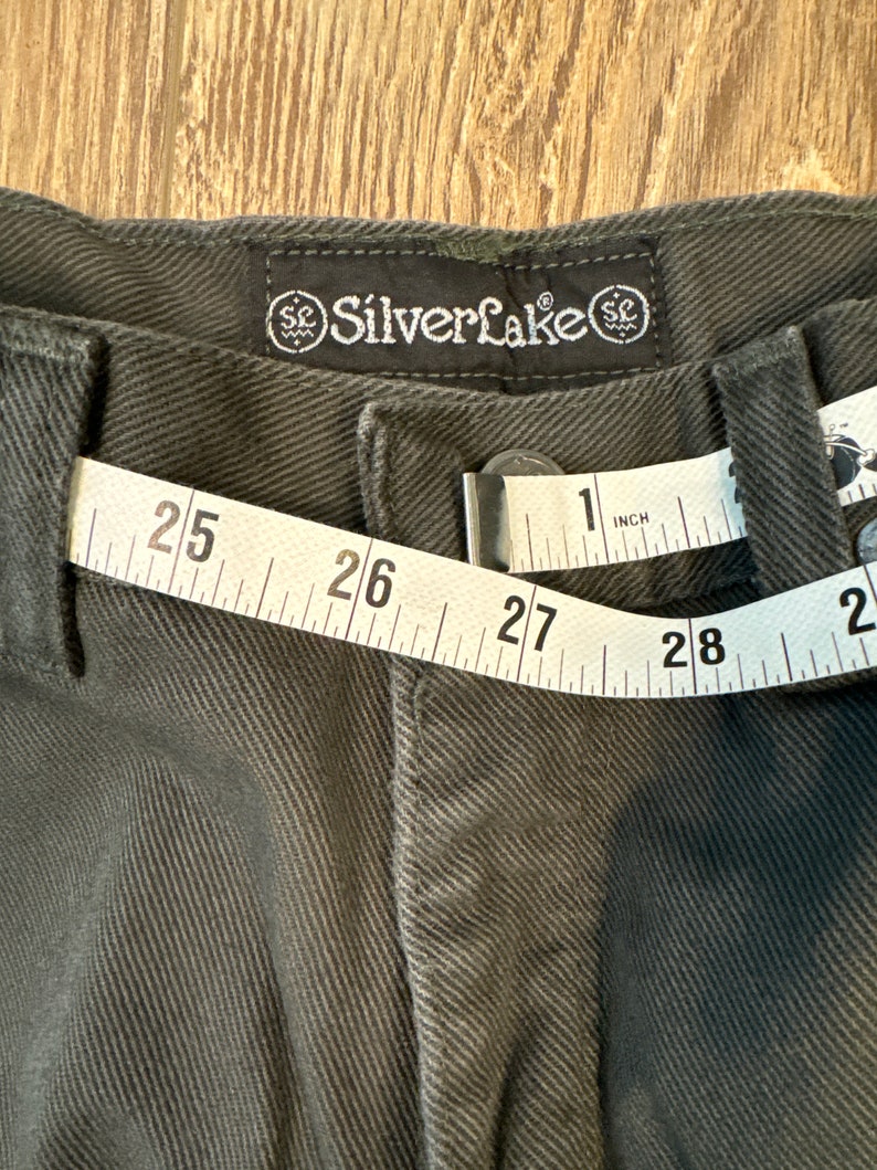 Op de afbeelding: Een donkergrijze broek met een meetlint om de taille. Het lint geeft 25 tot 28 inch aan. De broek heeft een label met de tekst "Silverlake" en een gestileerd "SC"-logo.